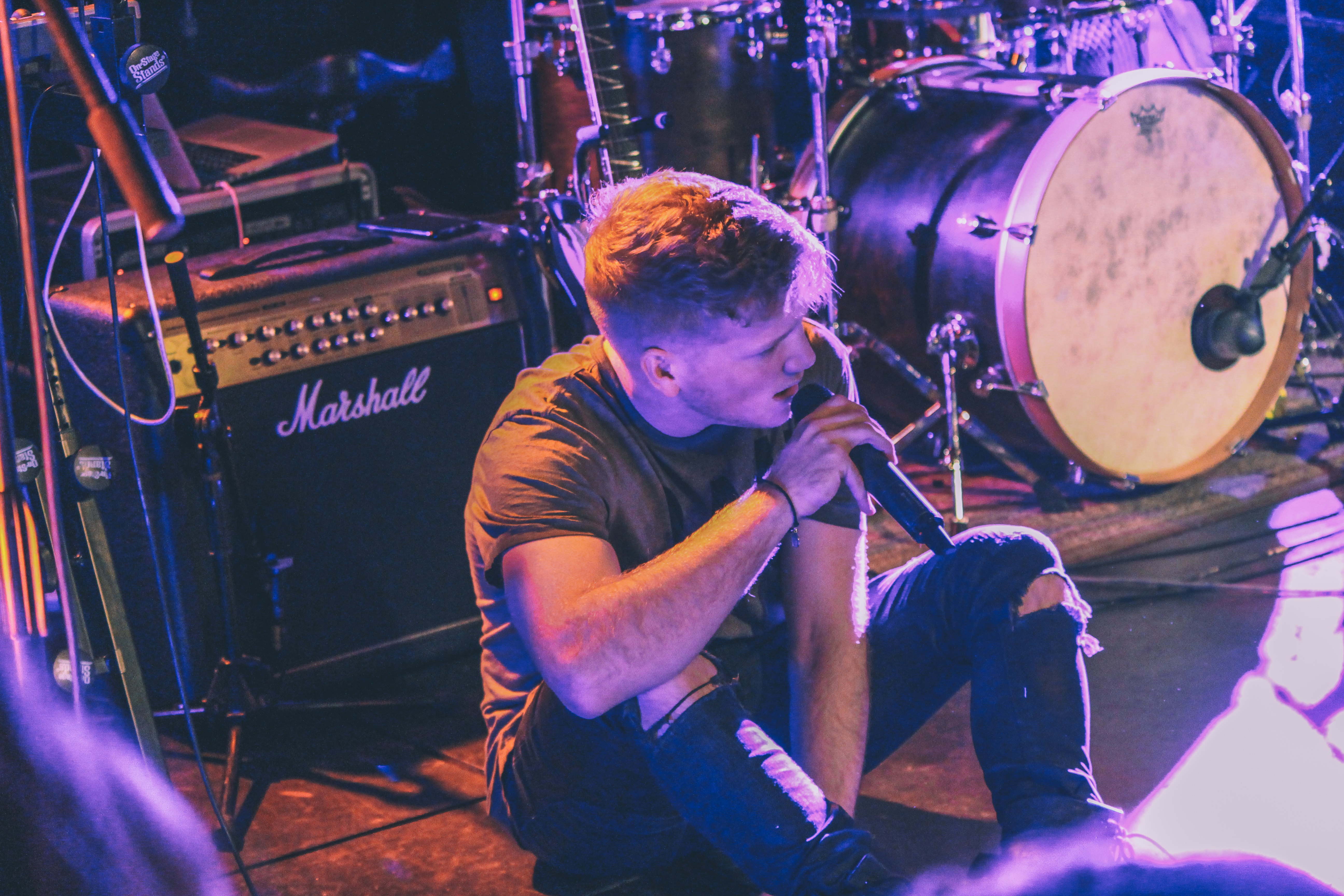 vocalist man holding microphone sitting in front of Marshall guitar amplifier near drum kit